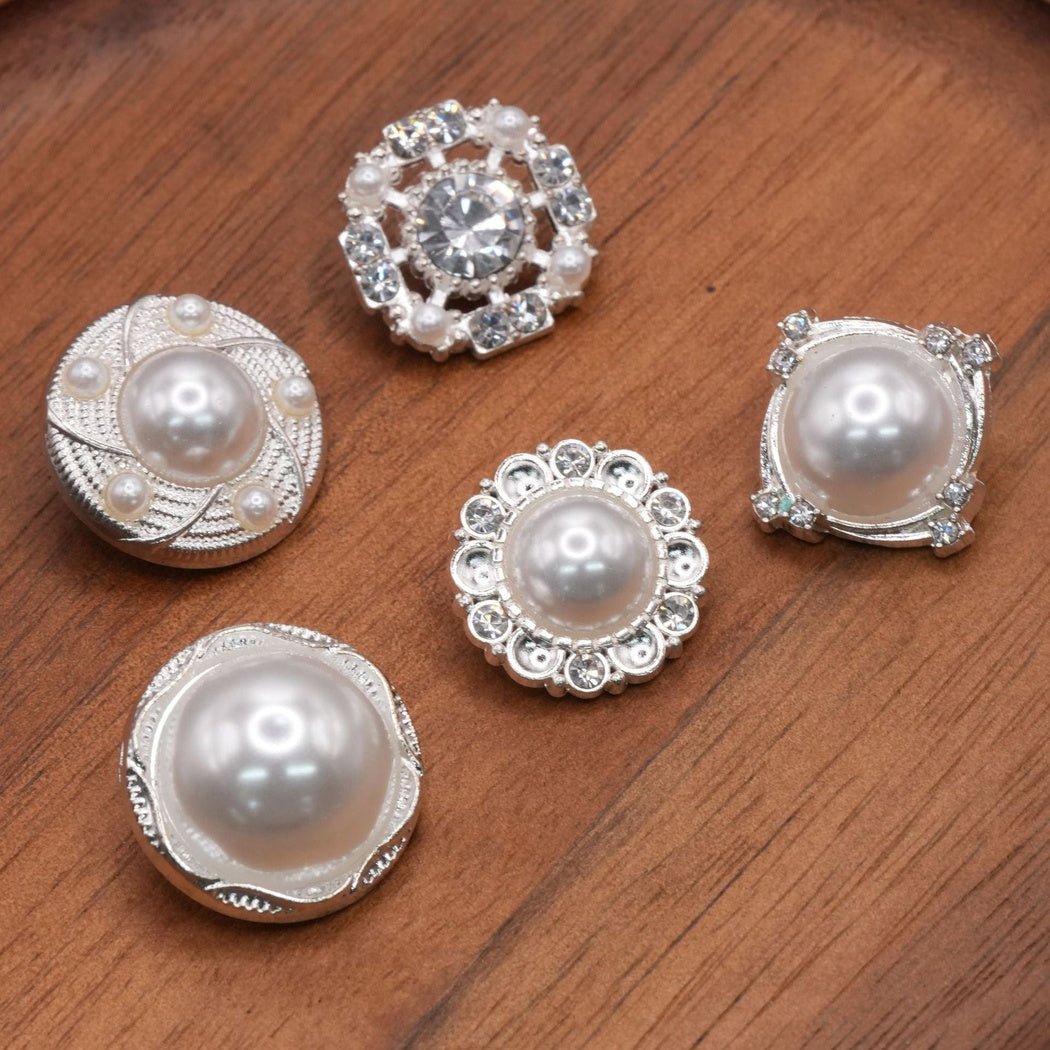 Five silver-tone decorative button designs with white domed or clear faceted centers on a brown surface.